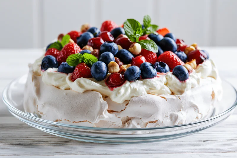 Pavlova meringue cake topped with fresh berries and cream on a glass plate on a white wooden table