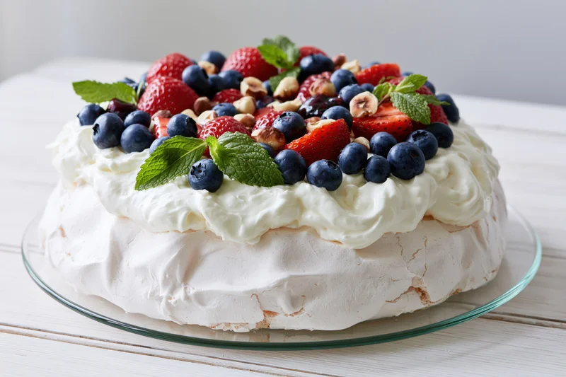 Pavlova meringue cake topped with blueberries strawberries cream and mint on a glass plate on a white wooden table