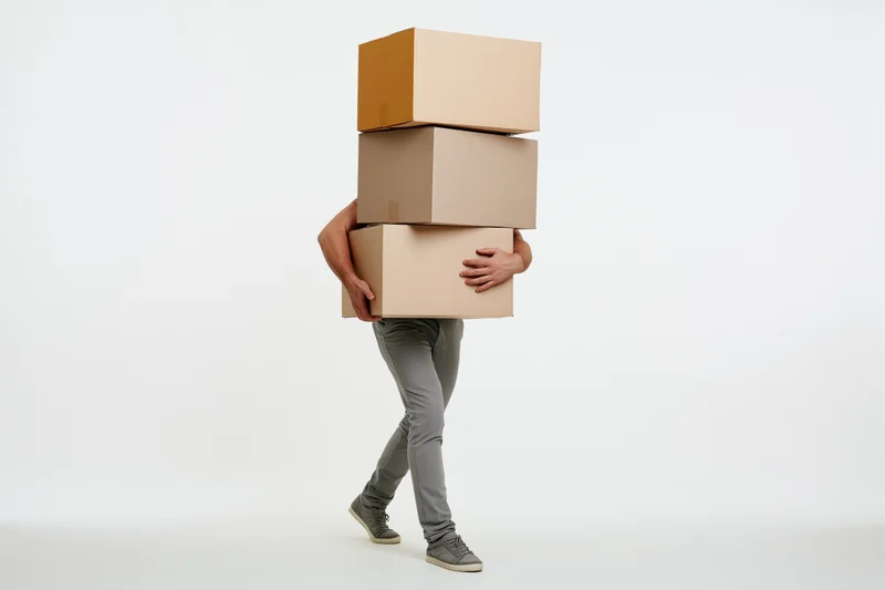 Man carrying a tall unstable stack of four large cardboard boxes in a studio setting