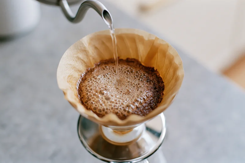 Water pouring from a kettle into a paper filter holding bloomed coffee grounds over a glass carafe on a countertop