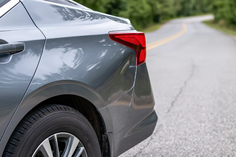 Rear quarter panel of a grey sedan with a dented fender on a curving road