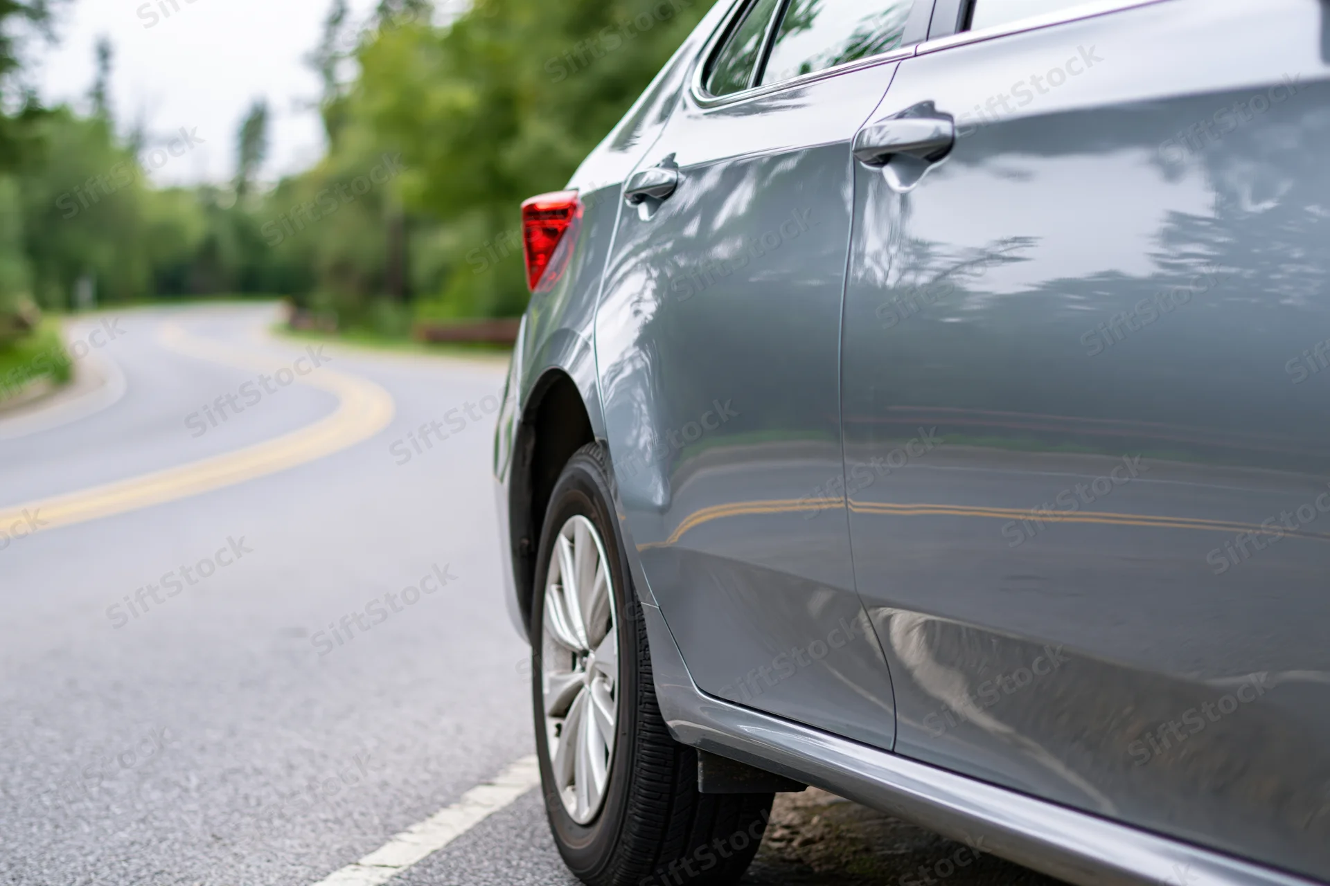 Grey sedan with dented rear fender parked on curved asphalt road beside green trees