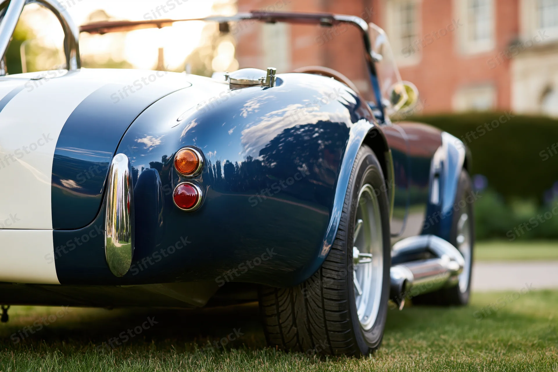 Rear quarter view of a blue and white classic ac cobra roadster parked on grass with sunlight reflecting on its glossy body