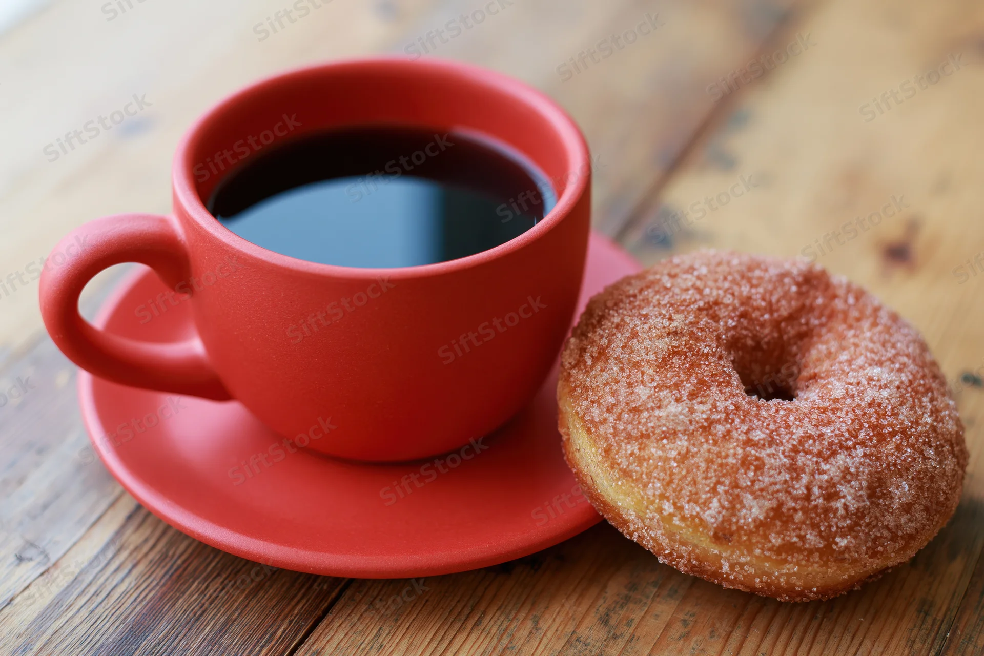 Red ceramic coffee cup with black coffee and sugar-coated donut on wooden table