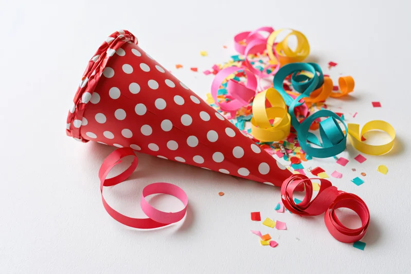 Red polka-dot party cone popper lying on its side with colorful streamers and confetti scattered around on white background