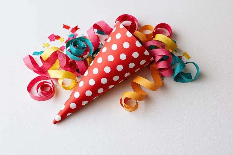 Red polka-dot party cone popper lying on its side with colorful streamers and confetti on white surface