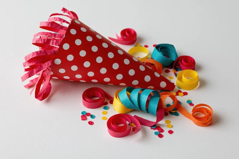 Red polka-dot party cone popper lying on its side with colorful streamers and confetti scattered around on white surface