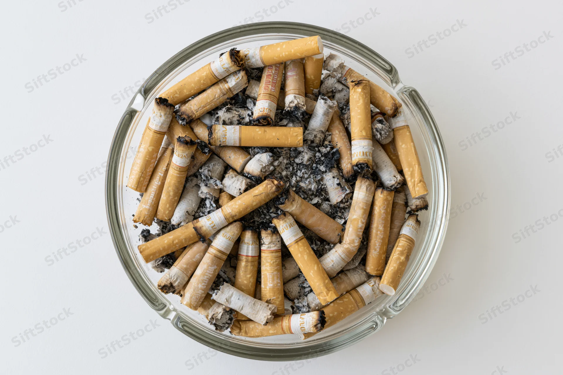 Round glass ashtray filled with cigarette butts and ash on white surface