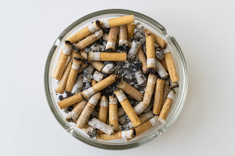 Round glass ashtray filled with cigarette butts and ash on white surface