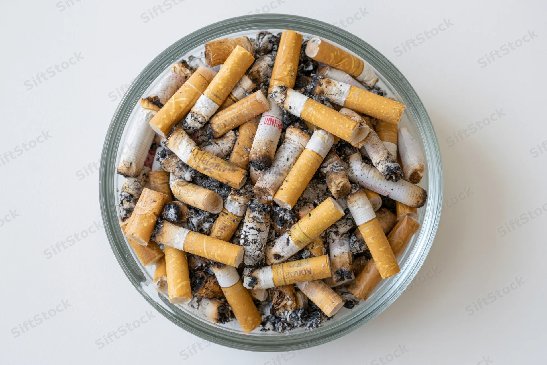 Round glass ashtray filled with cigarette butts and ash on white surface