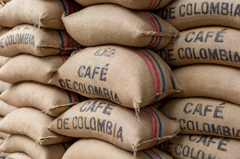 Stacked burlap coffee sacks labeled cafe de colombia in a warehouse