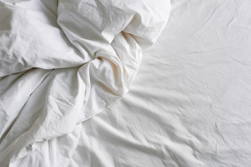 Rumpled white bedsheet and duvet bunched toward the upper-left on an unmade bed