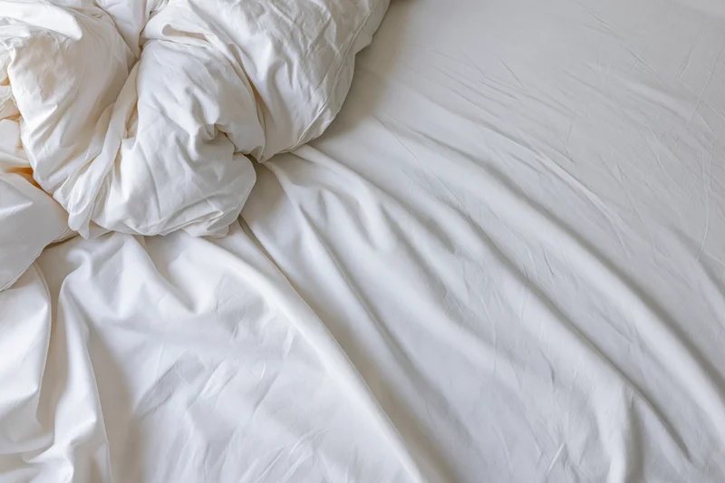Rumpled white bedsheet and duvet bunched toward the upper-left corner of an unmade bed