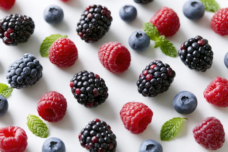 Scattered mix of fresh blackberries raspberries and blueberries with mint leaves on white surface