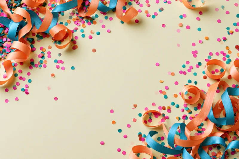 Colorful paper confetti and curled ribbons arranged in corners on a beige surface with copyspace in center