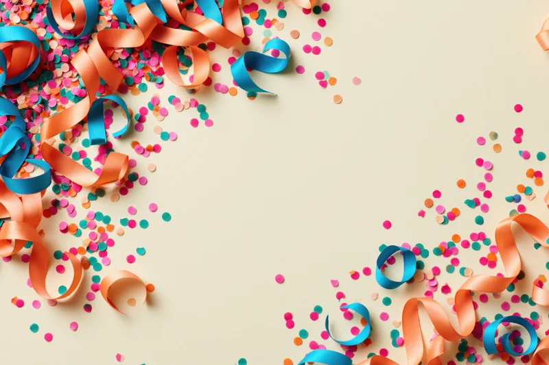 Multicolored paper confetti and curled ribbons scattered on beige surface with copyspace for party decoration