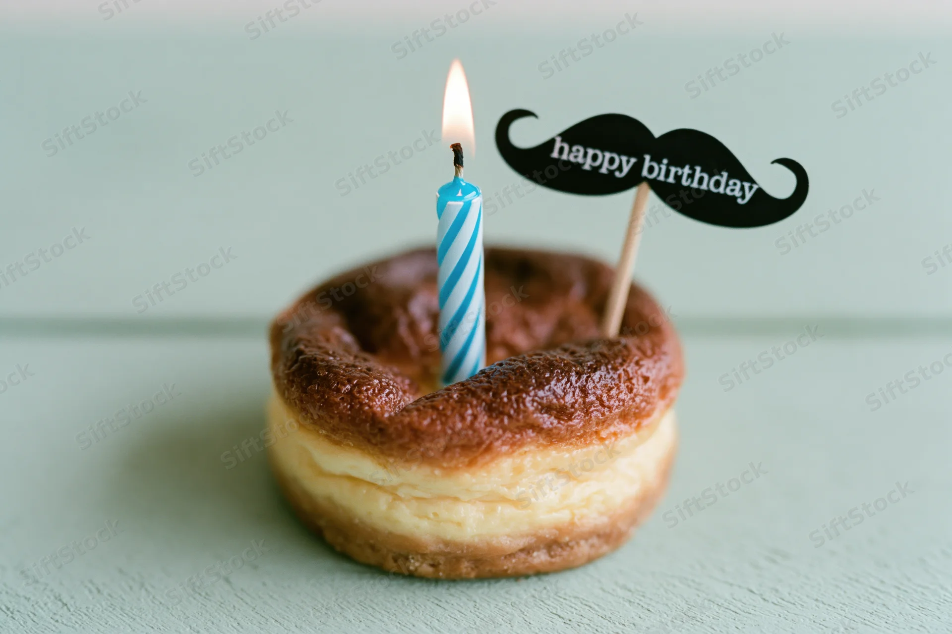Small round baked cheesecake with lit blue and white striped candle and mustache-shaped happy birthday sign on mint green surface