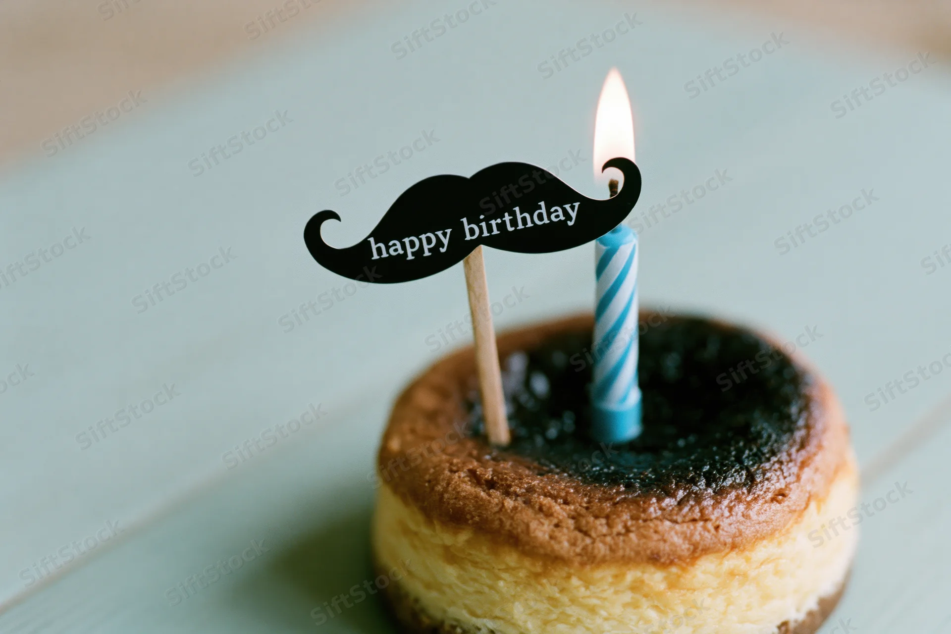 Small round cheesecake with lit blue and white striped candle and mustache topper on wooden surface