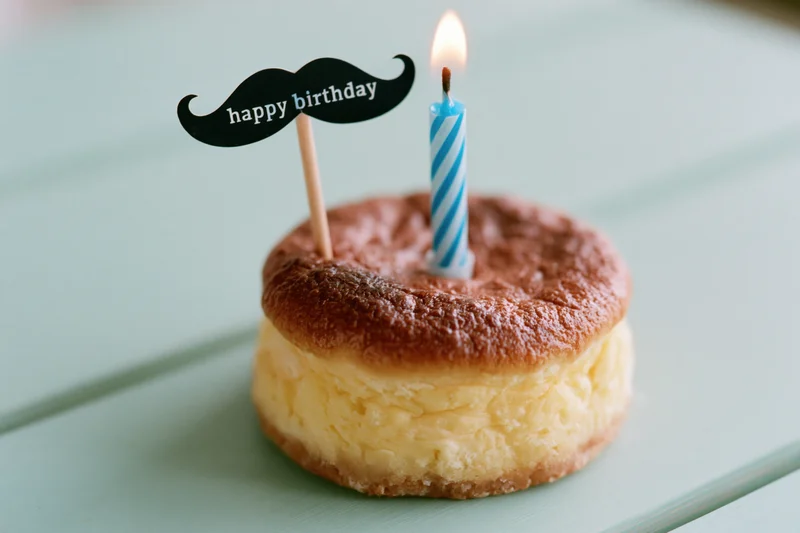 Small round cheesecake with lit blue striped candle and mustache-shaped happy birthday topper on mint green wooden surface