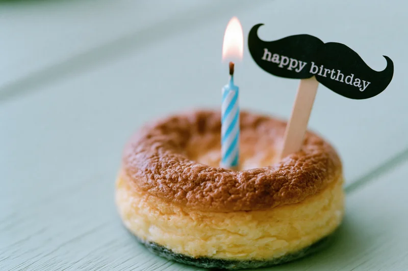 Small round baked cheesecake with lit blue and white striped candle and mustache-shaped happy birthday topper on wooden surface