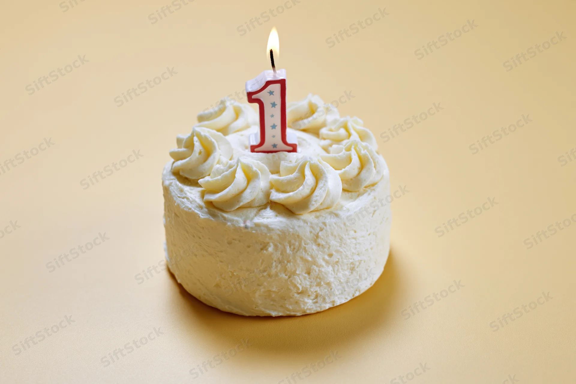 Small round white frosted cake with a lit number one candle on a beige surface
