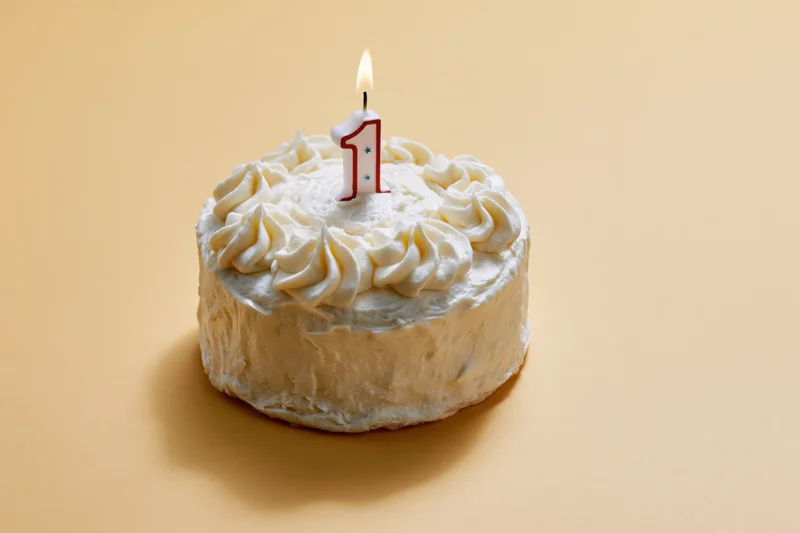 Small round white frosted cake with a lit number one candle on beige background