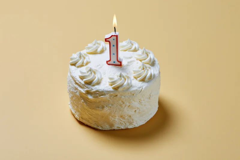 Small round white frosted cake with a lit number one candle on beige background