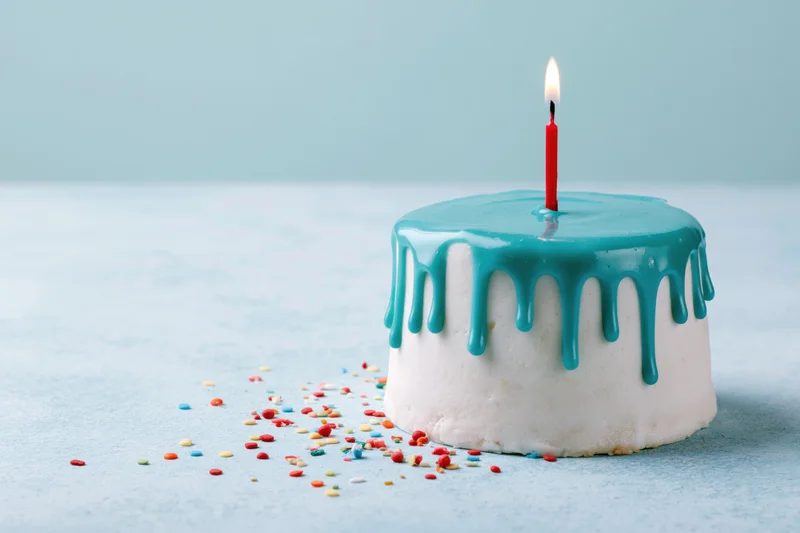 Small white birthday cake with teal drip frosting and one lit red candle on light blue surface with scattered sprinkles