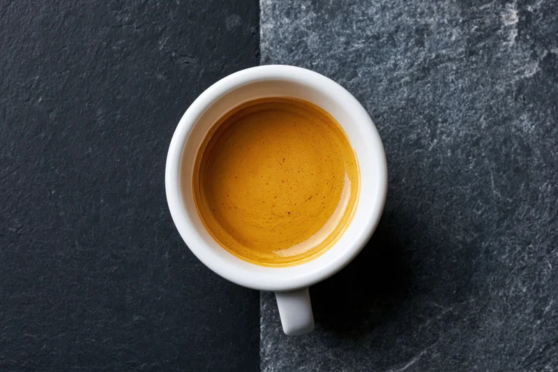 White ceramic espresso cup filled with golden-brown crema on dark stone surface, top view