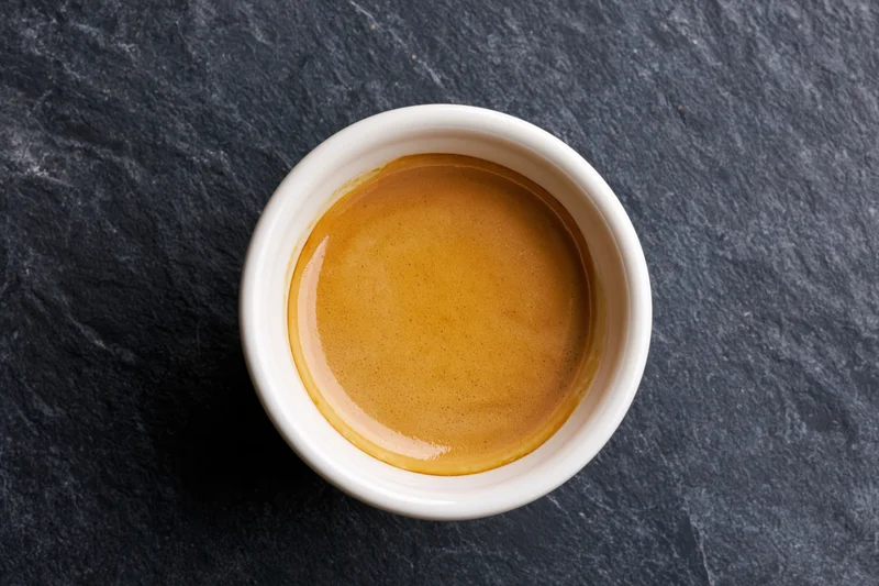 Overhead view of a white ceramic espresso cup filled with golden-brown crema on a dark slate surface
