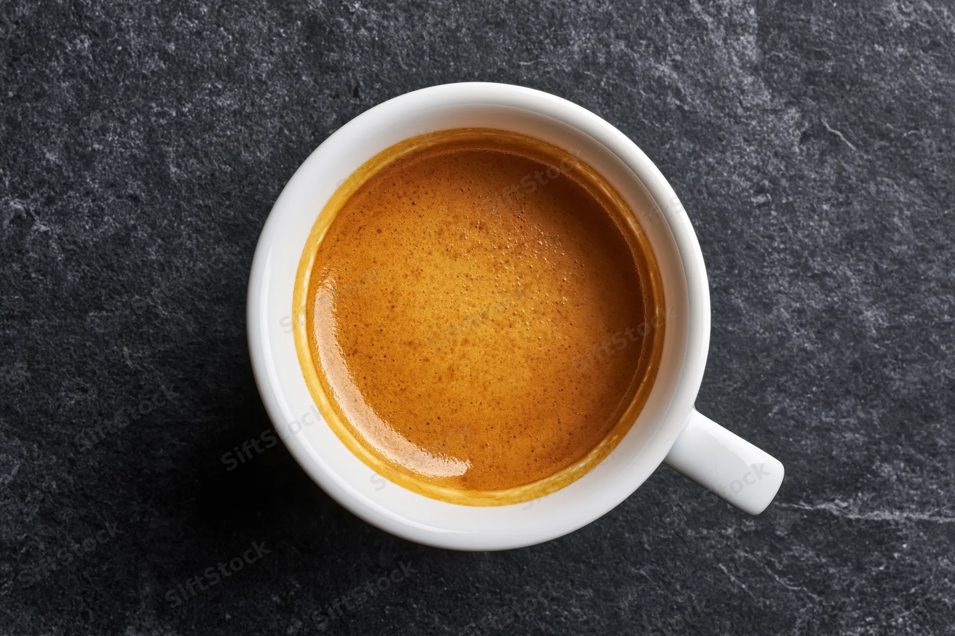 White ceramic espresso cup filled with golden-brown crema on dark stone surface, top view