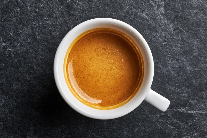 White ceramic espresso cup filled with golden-brown crema on dark stone surface, top view