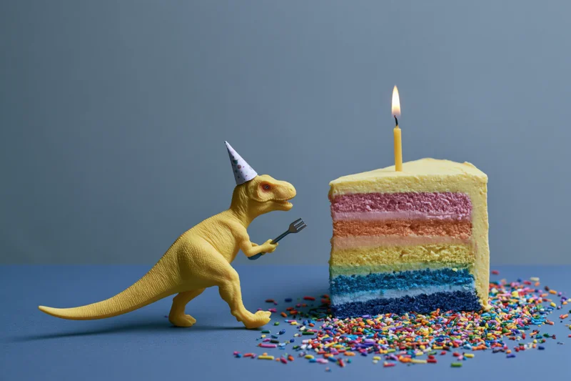 Yellow toy dinosaur wearing party hat and holding fork beside rainbow-layered birthday cake with lit candle and sprinkles