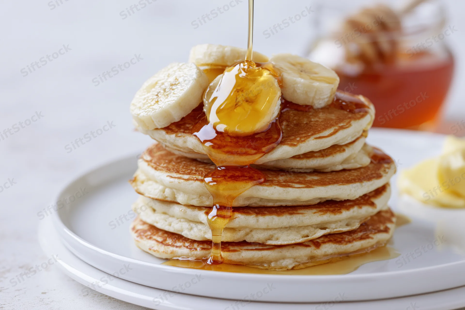 Stack of fluffy pancakes topped with banana slices and drizzling honey on a white plate