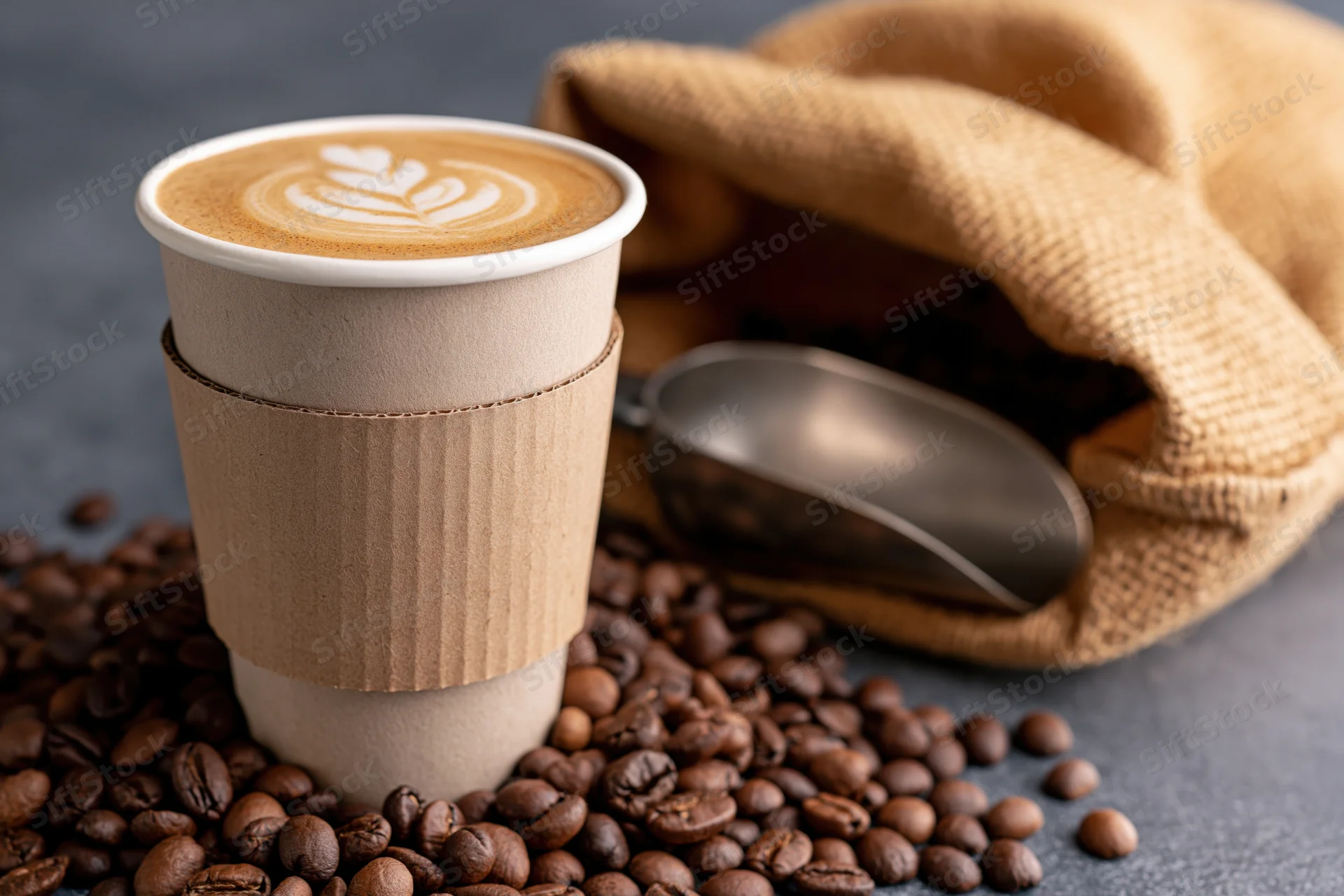 Takeaway paper cup with latte art and cardboard sleeve surrounded by coffee beans and a burlap sack with a metal scoop
