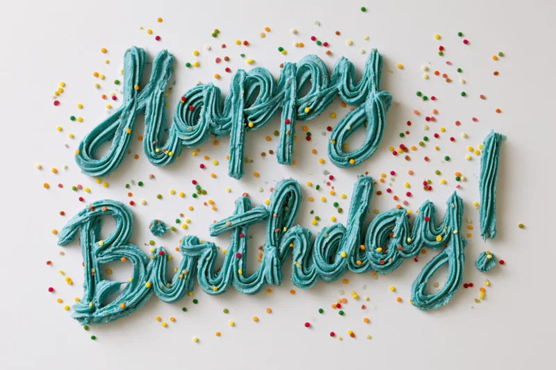 Teal buttercream frosting spelling happy birthday on white surface with colorful sprinkles scattered around