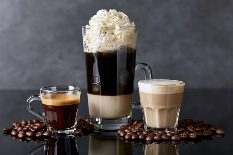 Three glass coffee drinks arranged on a reflective black surface with coffee beans scattered around