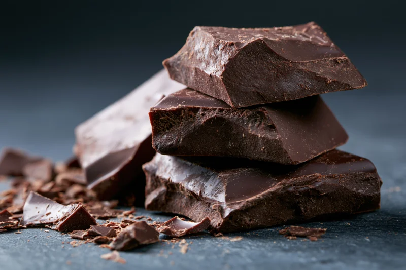 Broken dark chocolate chunks stacked unevenly on a dark surface with scattered crumbs