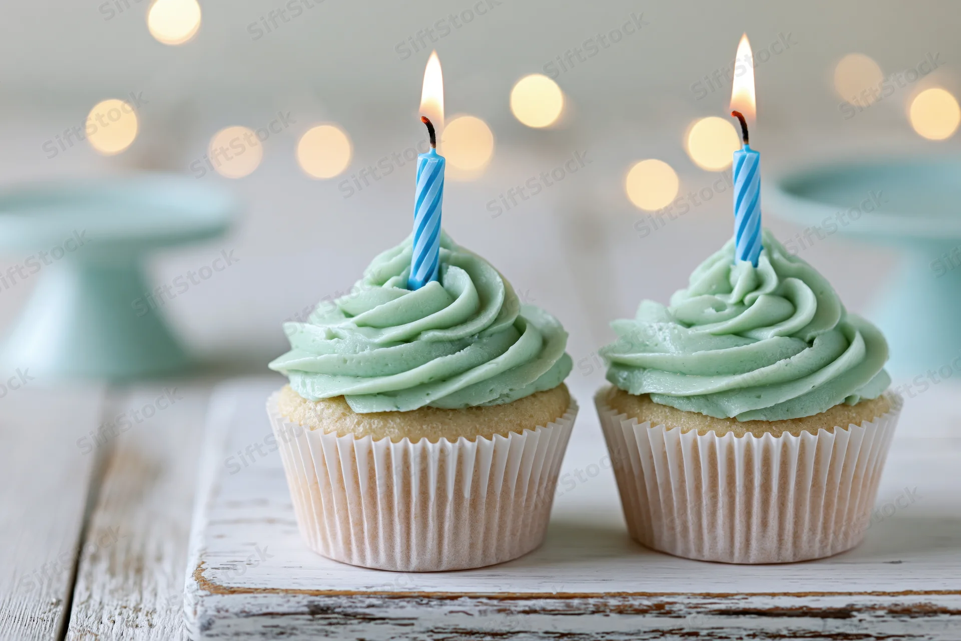 Two birthday cupcakes with swirled mint green frosting and lit candles on a rustic white wooden board
