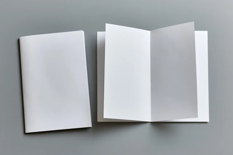 Two blank white paper booklets on a flat gray surface, one closed and one open, arranged side by side in a top-down view