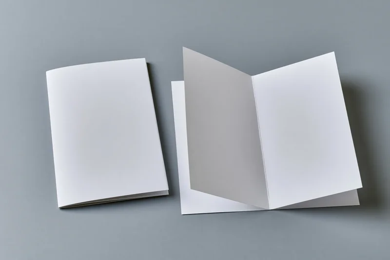 Two blank white paper booklets on a flat grey surface, one closed and one open, arranged side by side