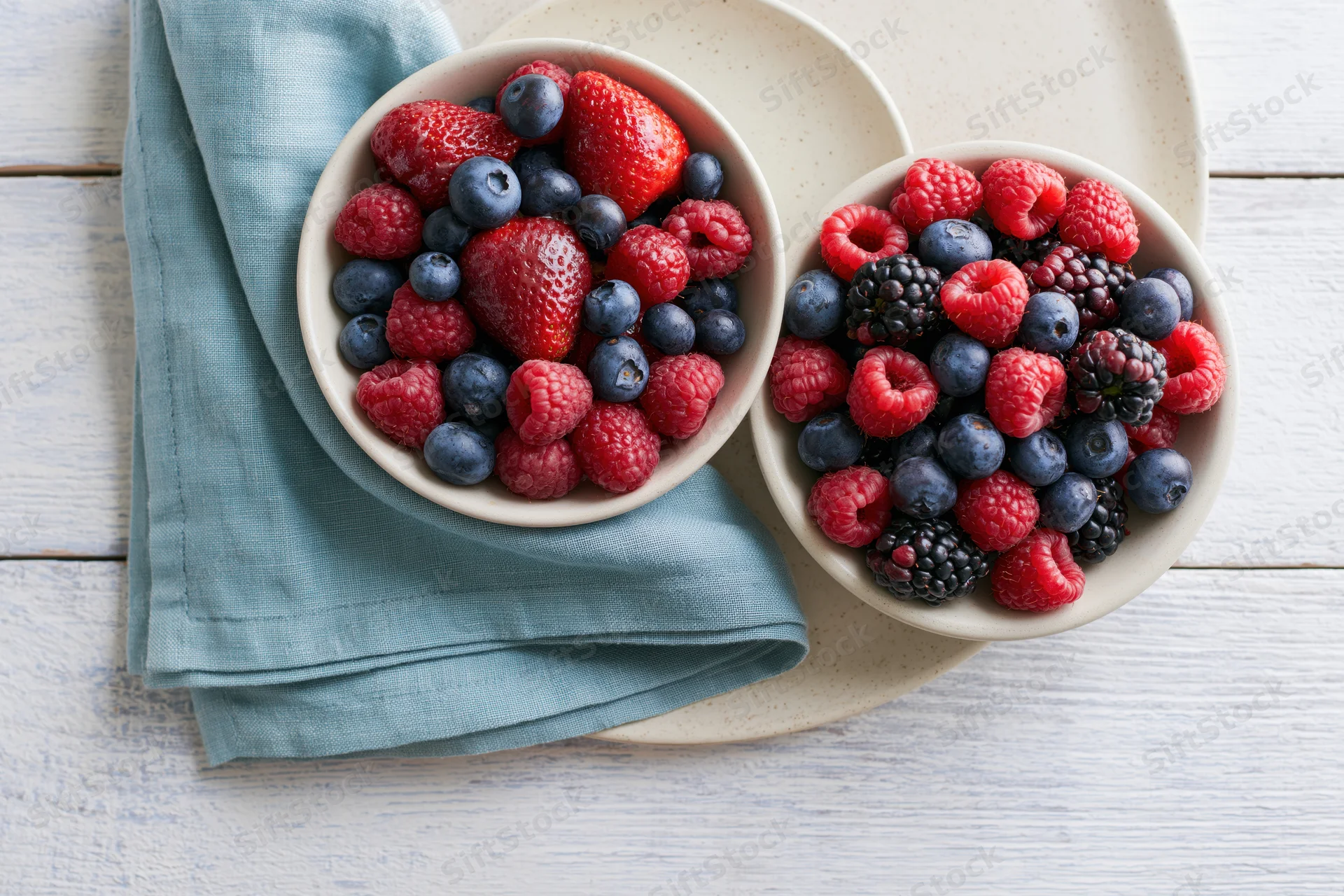 Two ceramic bowls filled with mixed fresh berries on a white wooden table with a blue napkin and plate