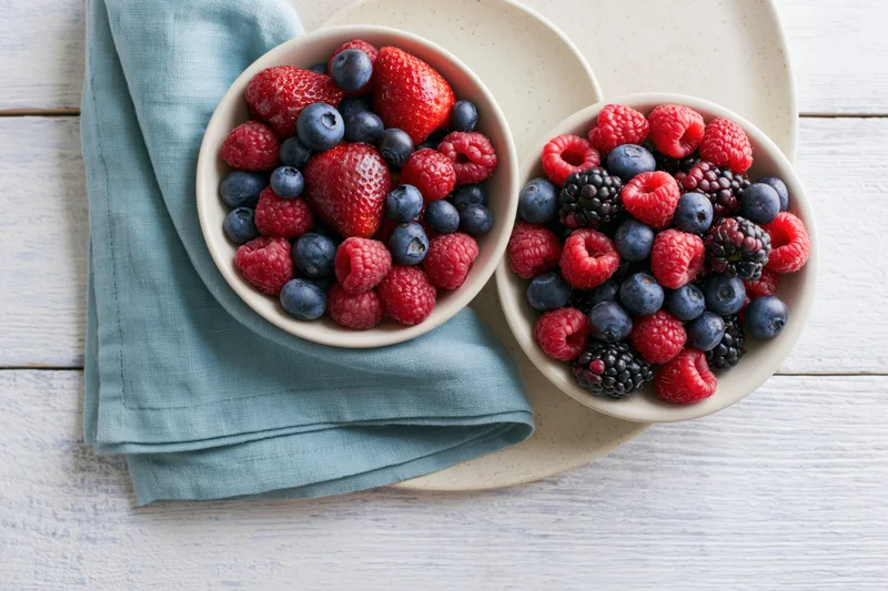 Two ceramic bowls filled with mixed fresh berries on a white wooden table with a blue napkin and plate