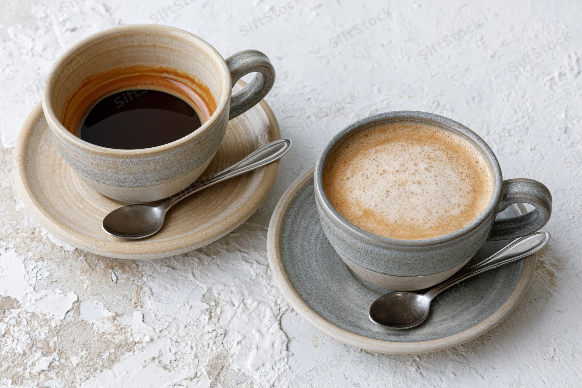 Two ceramic coffee cups with saucers and spoons on a textured white surface