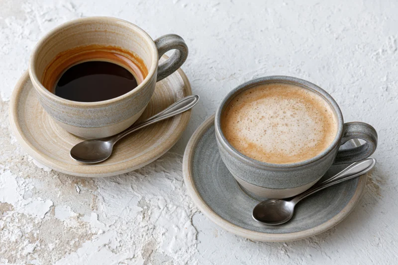 Two ceramic coffee cups with saucers and spoons on a textured white surface
