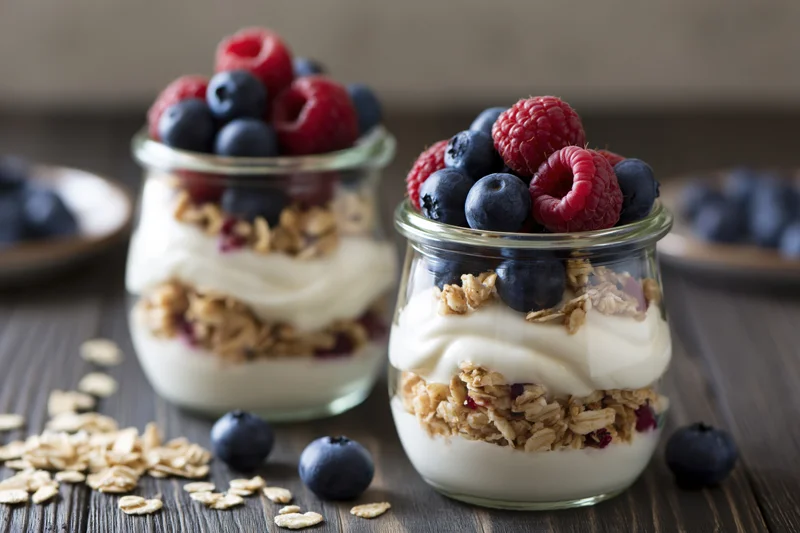Two glass jars layered with yogurt, granola and fresh berries on a dark wooden table