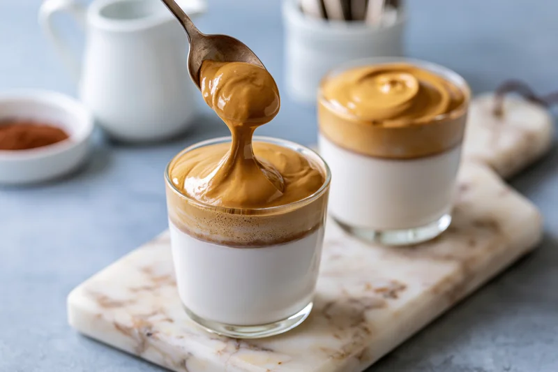 Spoon pouring caramel-colored whipped coffee topping onto a glass of milk-based drink on a marble serving board