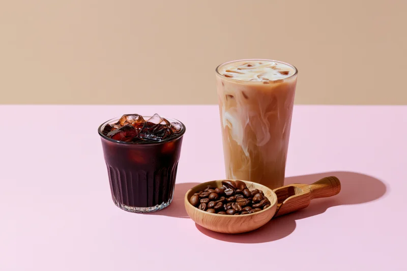 Two iced coffee drinks with coffee beans and wooden scoop on a pink surface