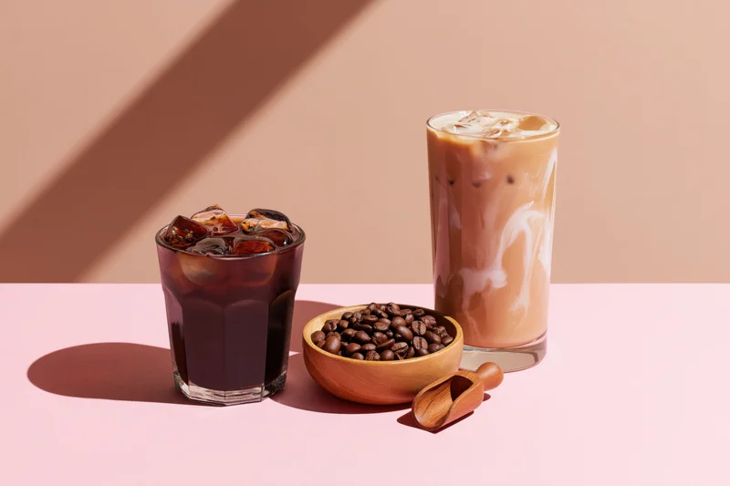 Two iced coffee drinks with coffee beans in a wooden bowl on a pink surface