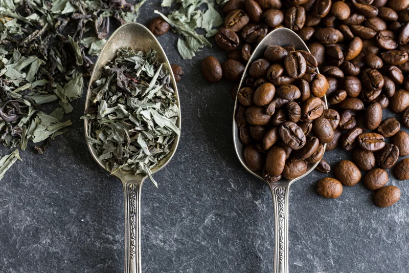 Two vintage silver spoons holding coffee beans and dried tea leaves on dark slate surface